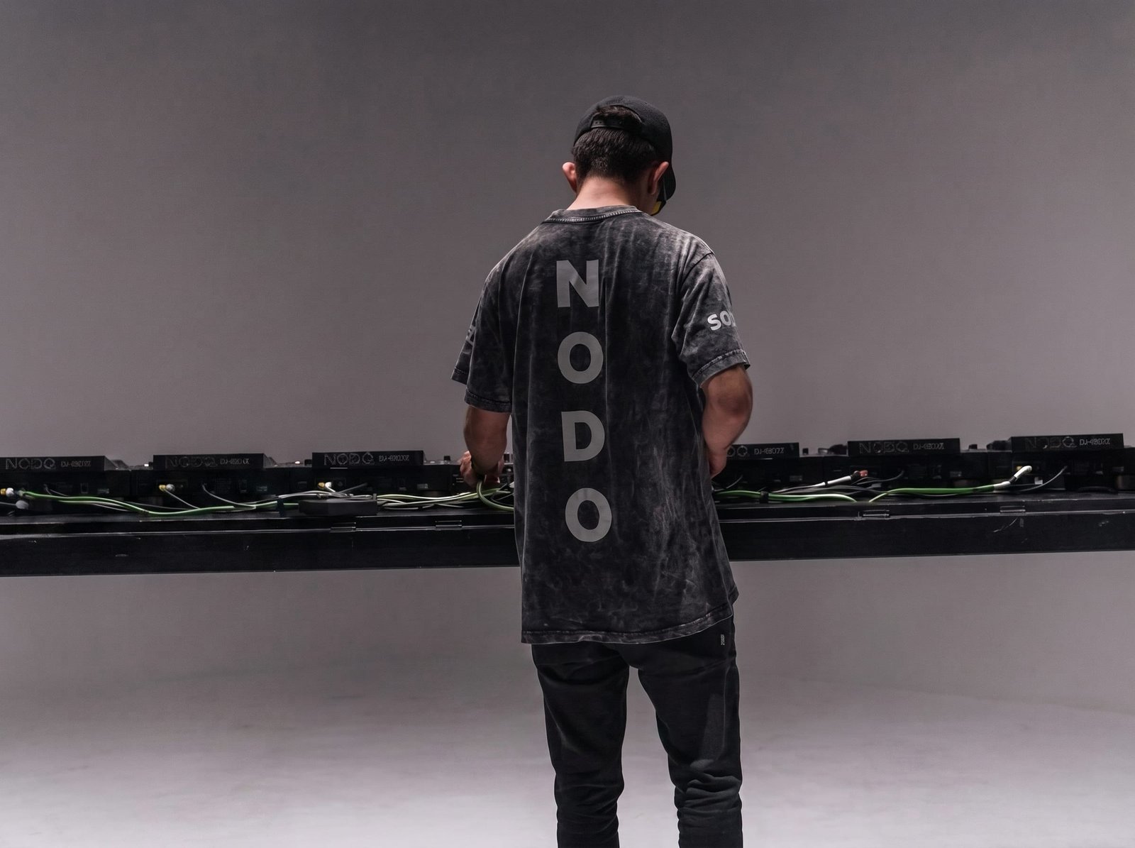 FerchX de espaldas frente a una fila de CDJs, playera Nodo